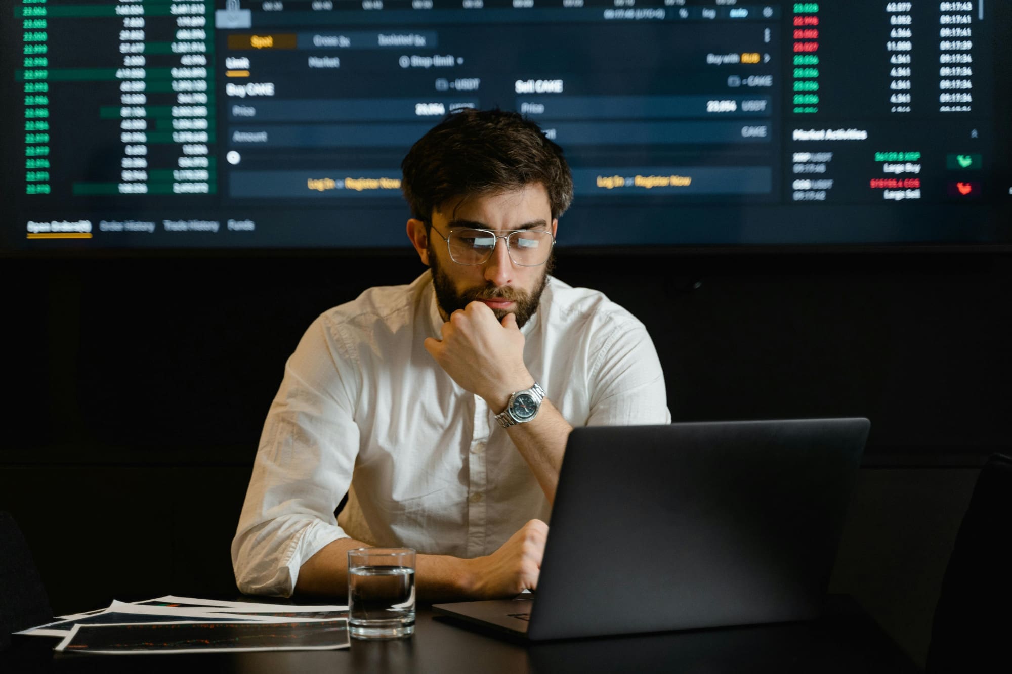 Office worker sitting at laptop looking confused with graphs on a screen behind him.