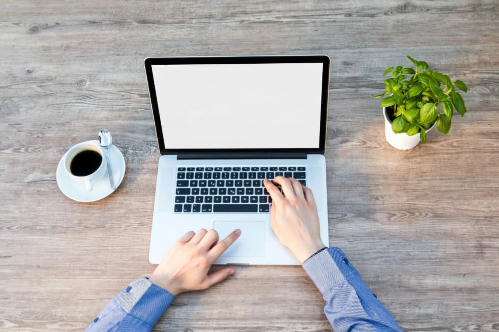 Person typing on laptop rested on wooden desk outside, between a cup of coffee and a desk plant.