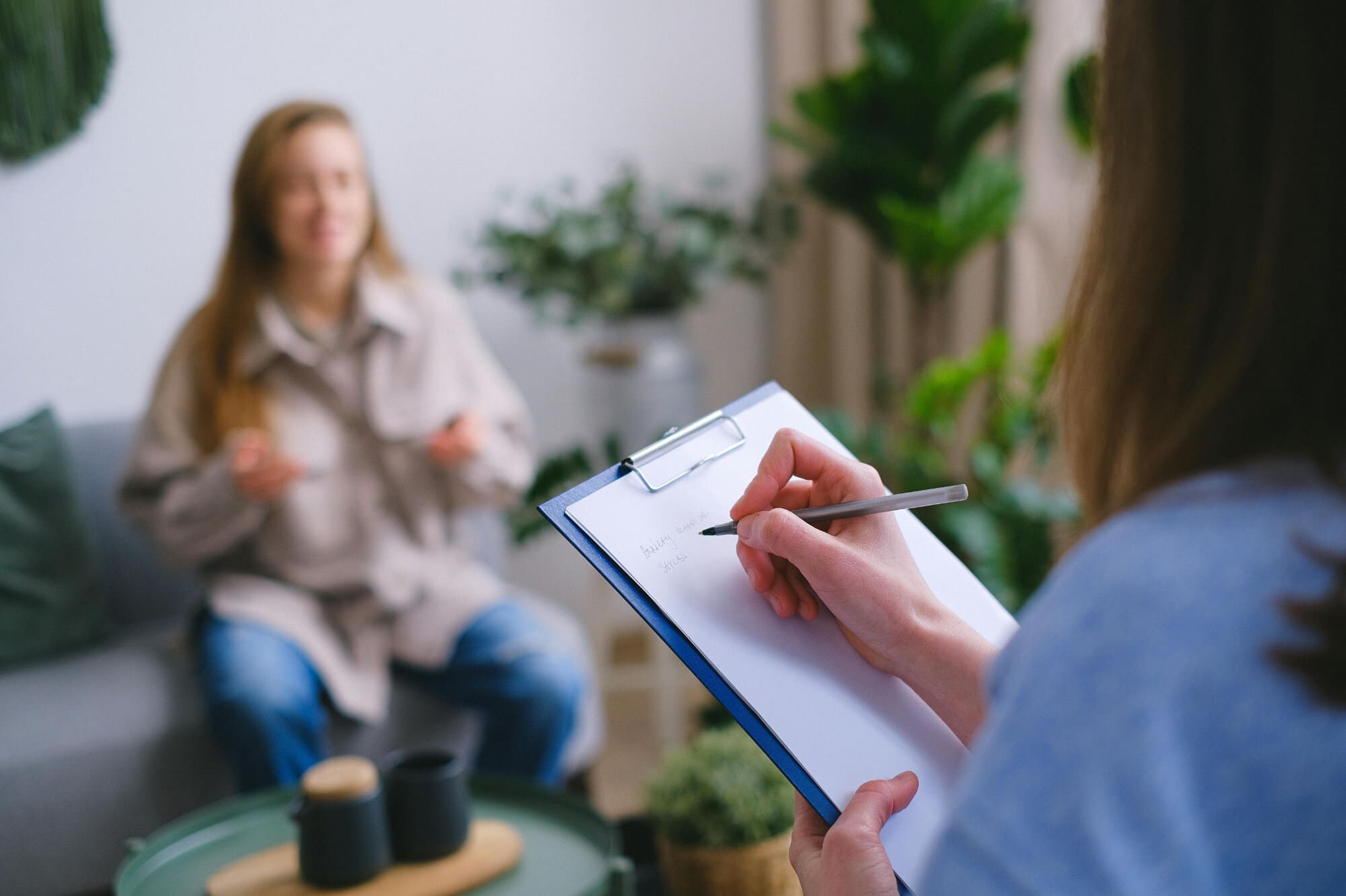 Woman writes on a notepad while sitting across from another woman sitting on a couch.