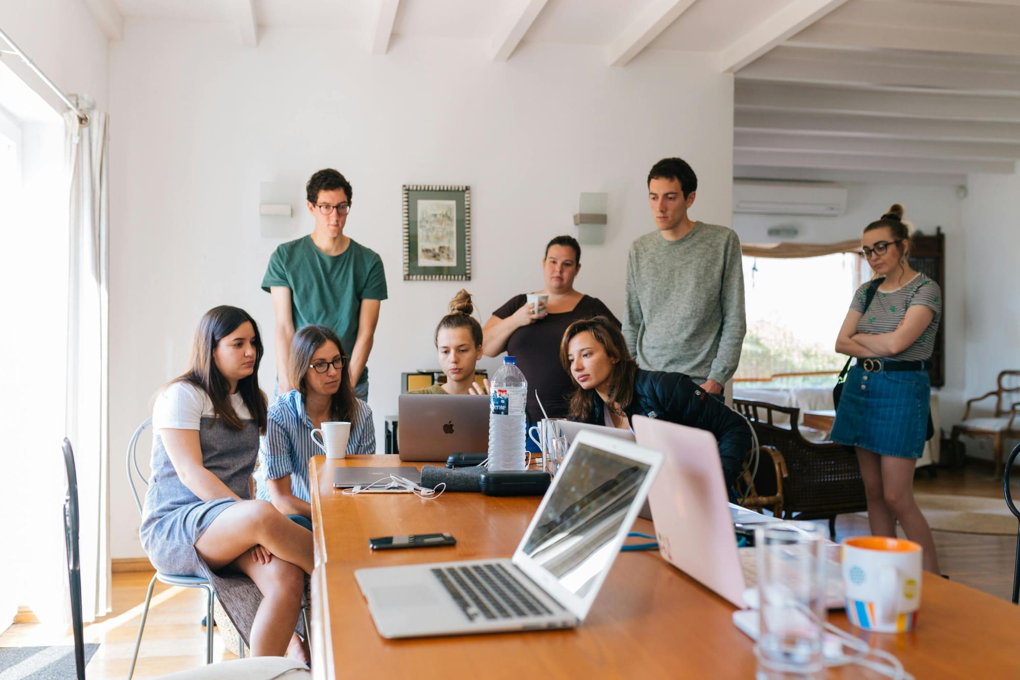 Several office workers standing and crouching around a laptop at the end of a long desk.