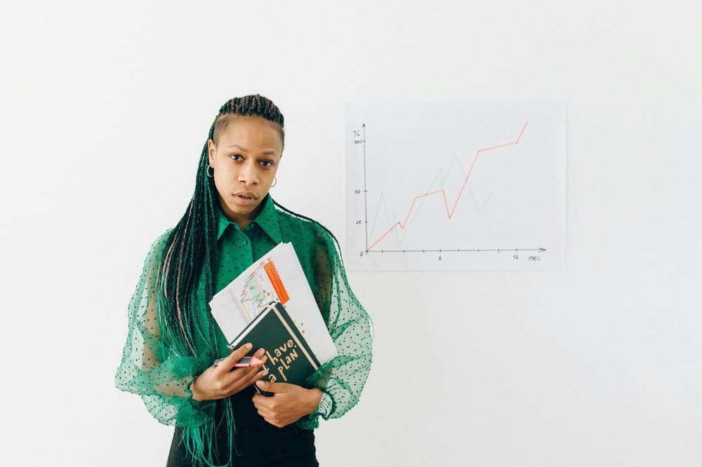 Office worker holding a notepad and documents while standing in front of whiteboard with business statistics written on it.