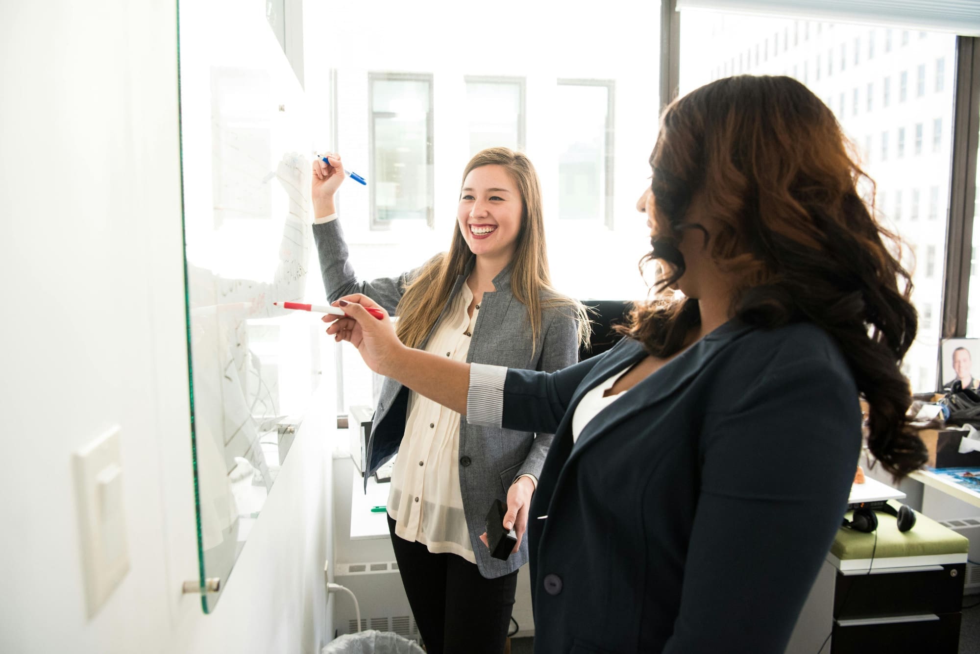 Two female office workers pointing whiteboard pens at a whiteboard and smiling.