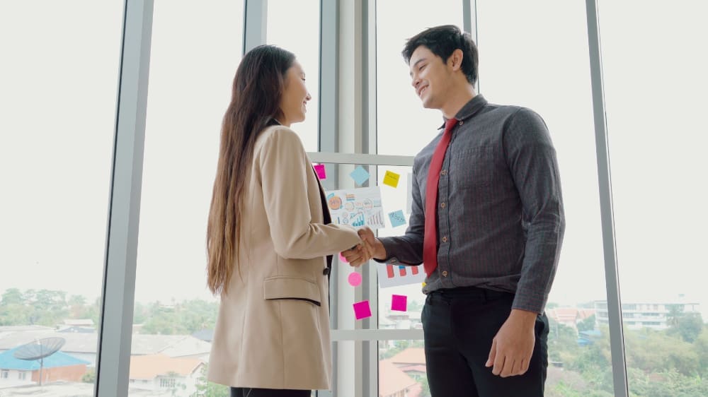 Two office workers shaking hands in front of whiteboard covered in post-it notes and business graphs.