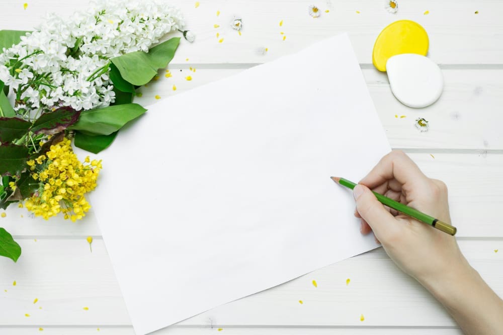 A hand holding green pencil up against blank sheet of paper on white desk next to green and yellow flowers.