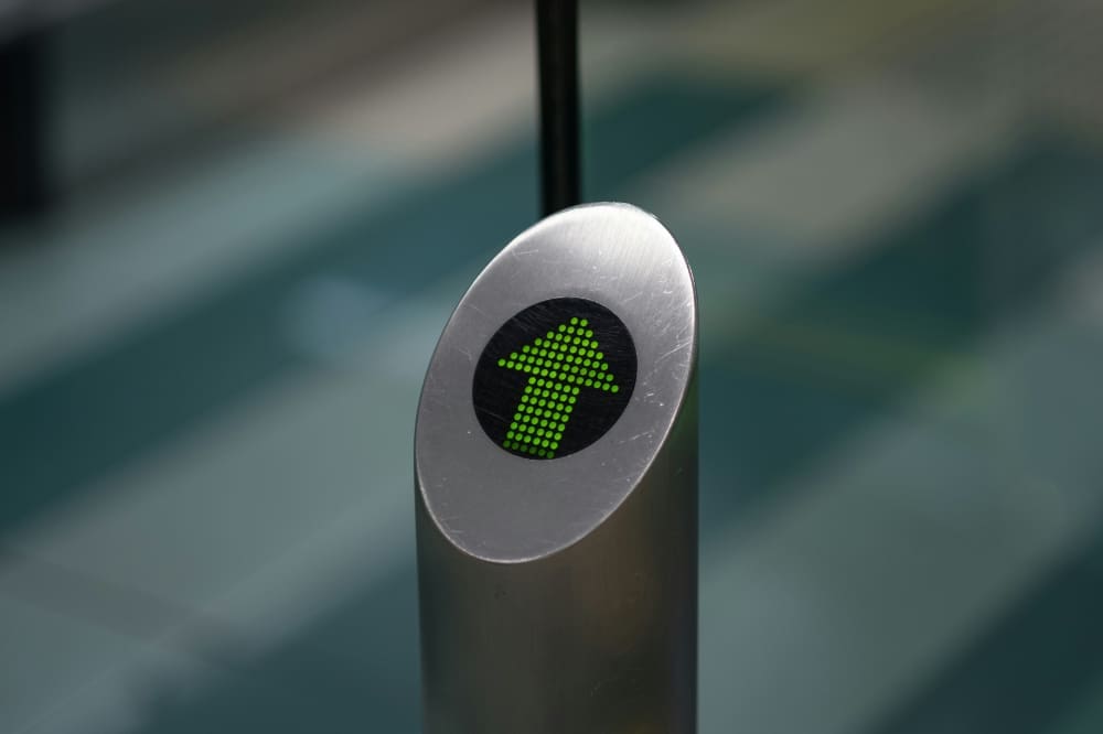 A silver metal street bollard with a screen portraying a green arrow pointing forwards.