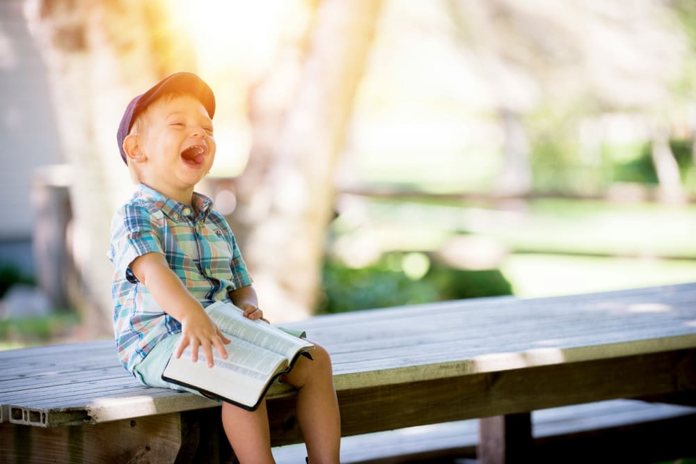 Young child sitting outside on wooden park table laughing with open book on his lap.