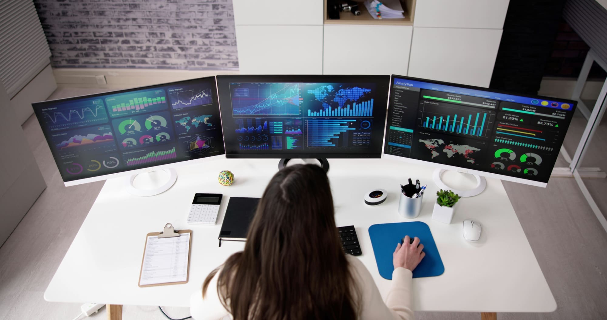Office worker sat at desk looking at three monitors side-by-side displaying data.