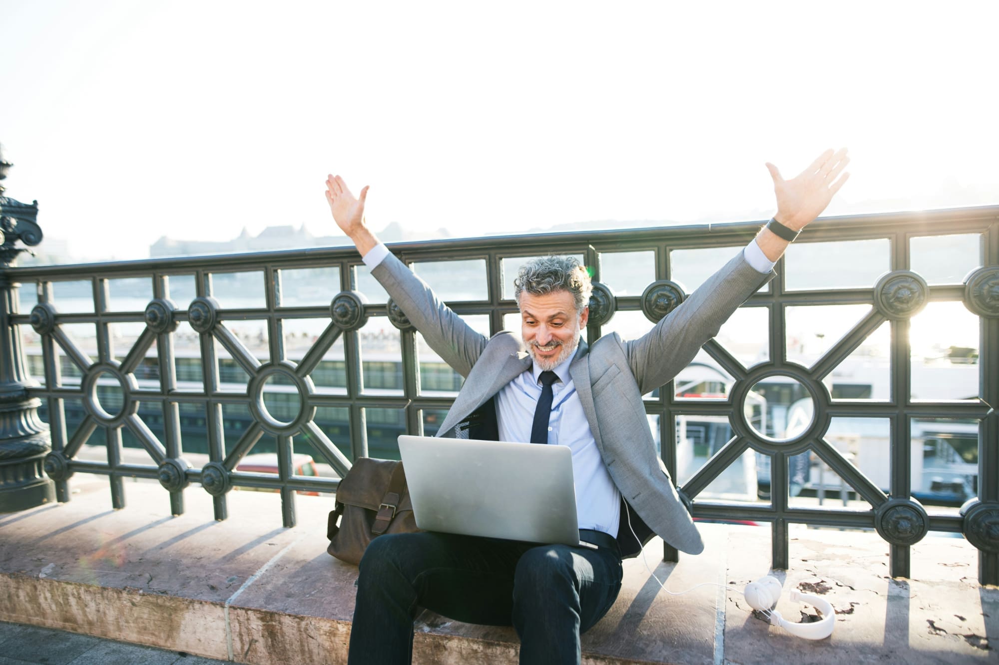 A man sitting on a bench outside celebrating in front of a laptop.