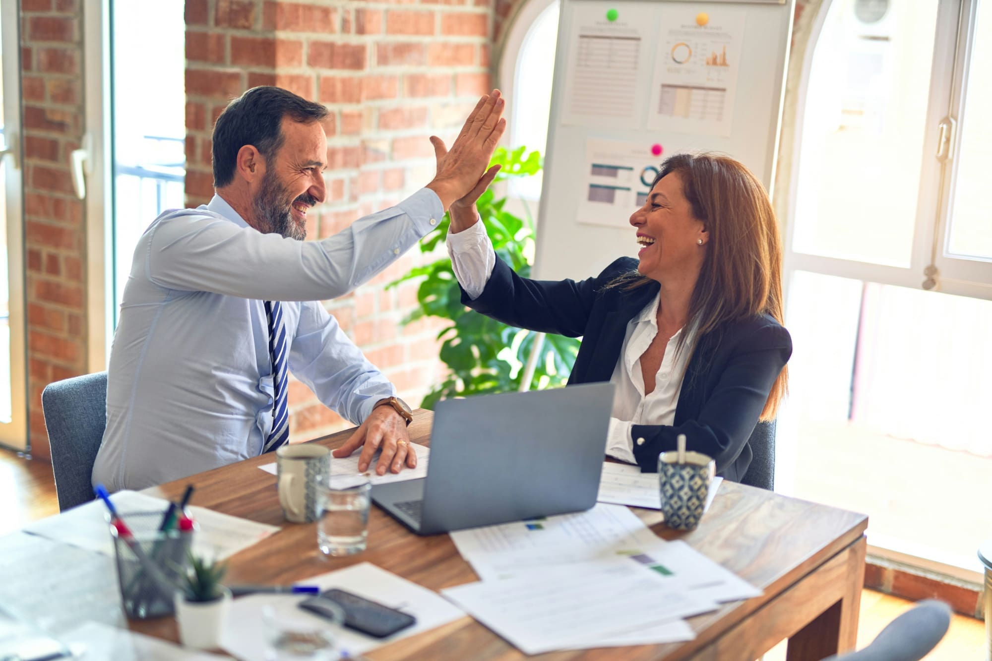 Two office workers high-fiving in front of desk with laptop and documents on it.