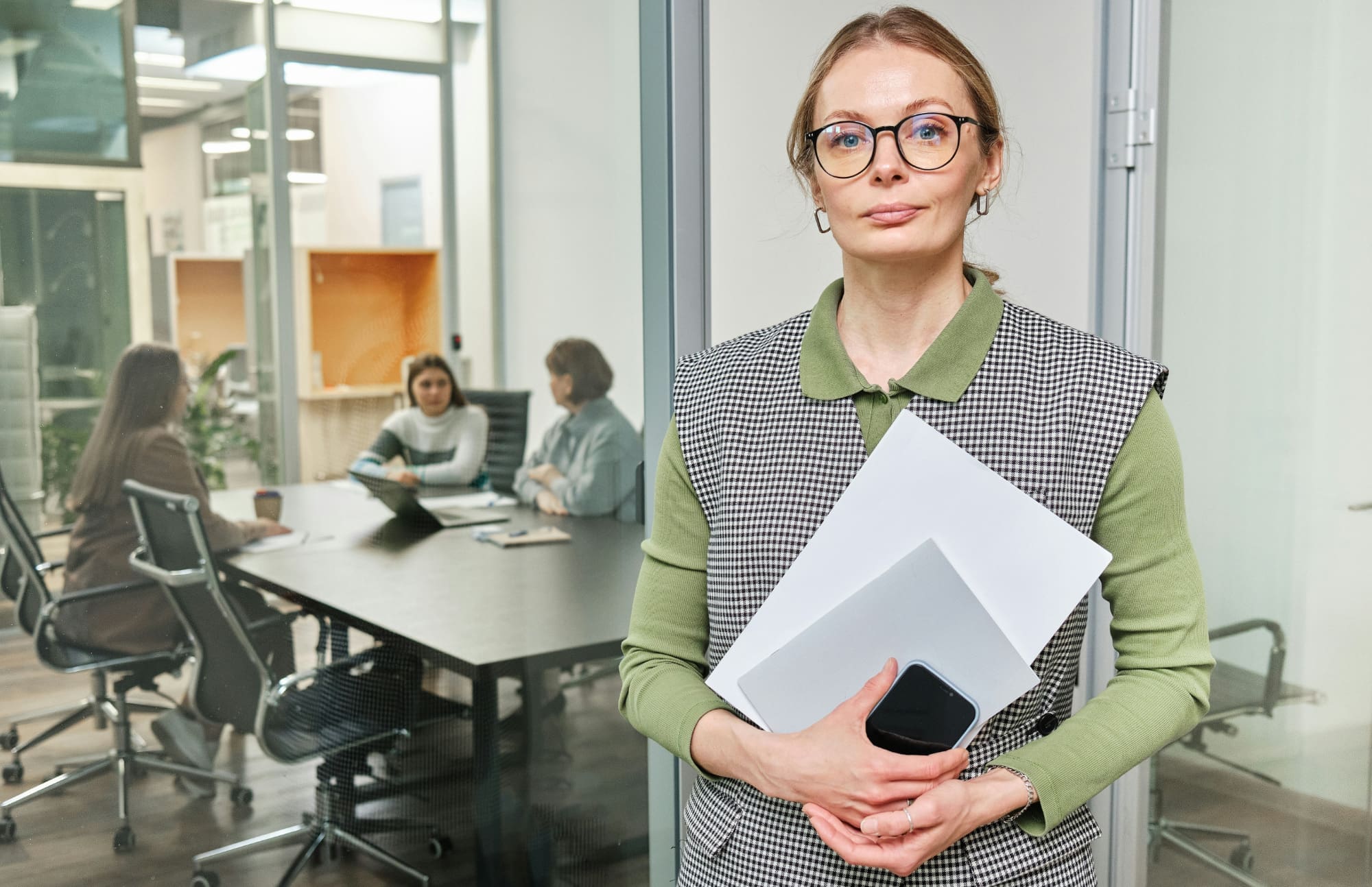Woman holding phone and documents in the office with colleagues sat at a table behind her.