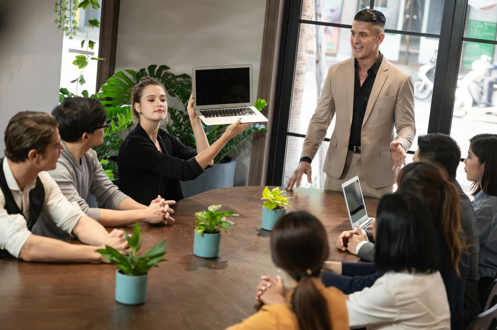 An office employee holding up a laptop as other employees around a table look at it.