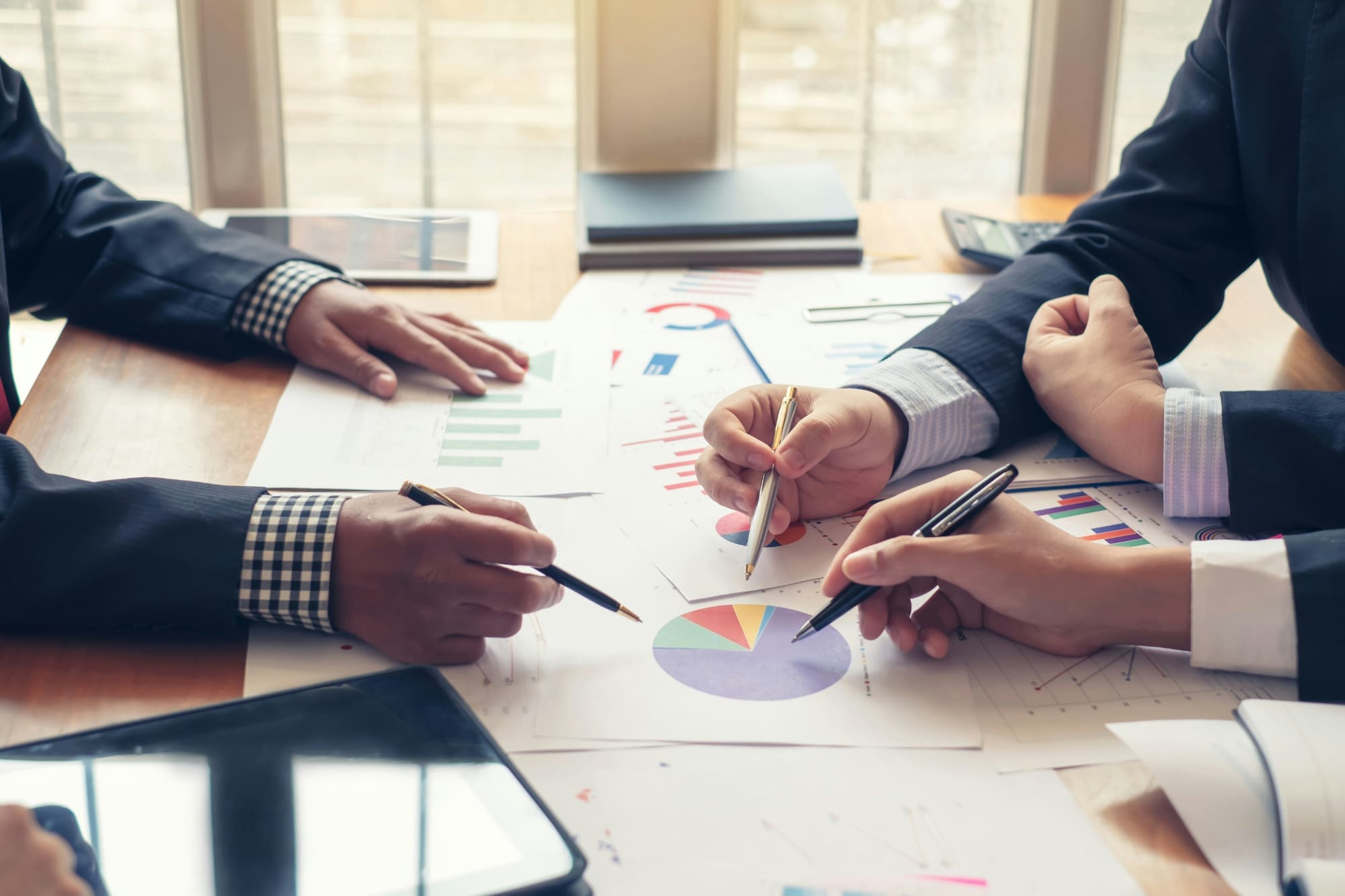 Multiple business professionals using pens to point at multiple graphs laid out on a table.