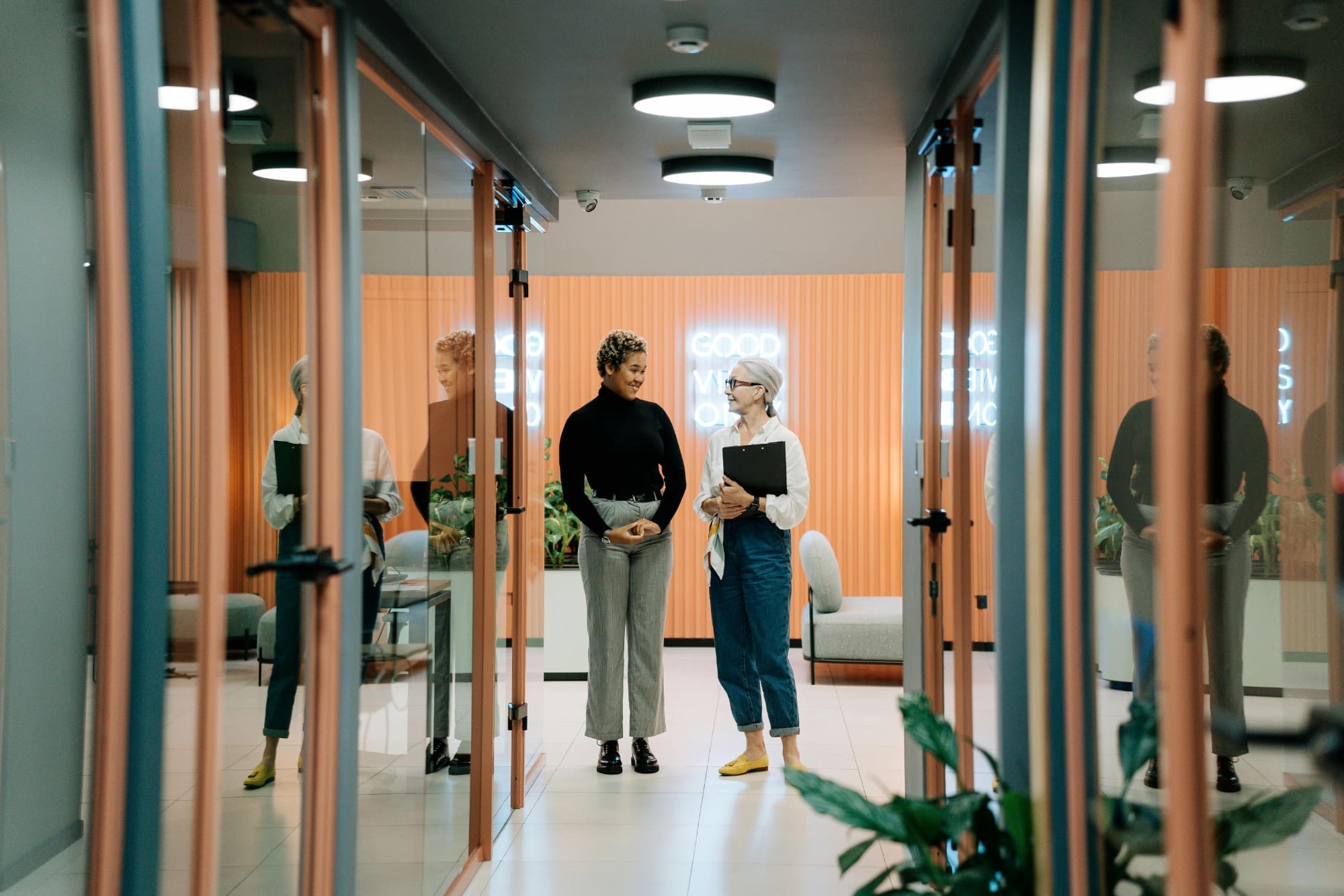 Two office workers standing next to each other in hallway talking and smiling.