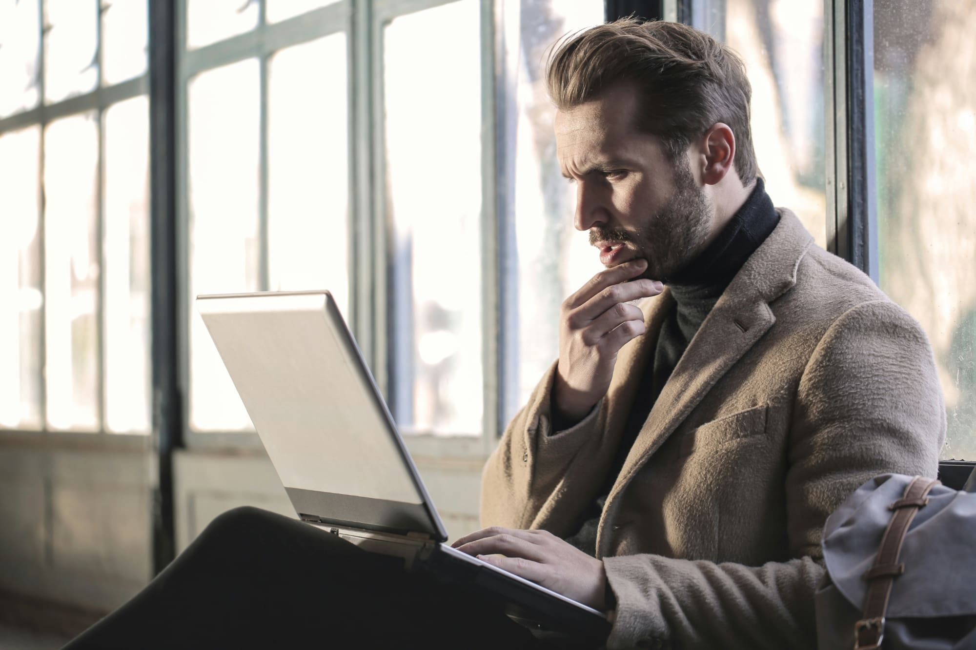A man sitting on a bench next to a window looking curiously at the laptop on his lap.