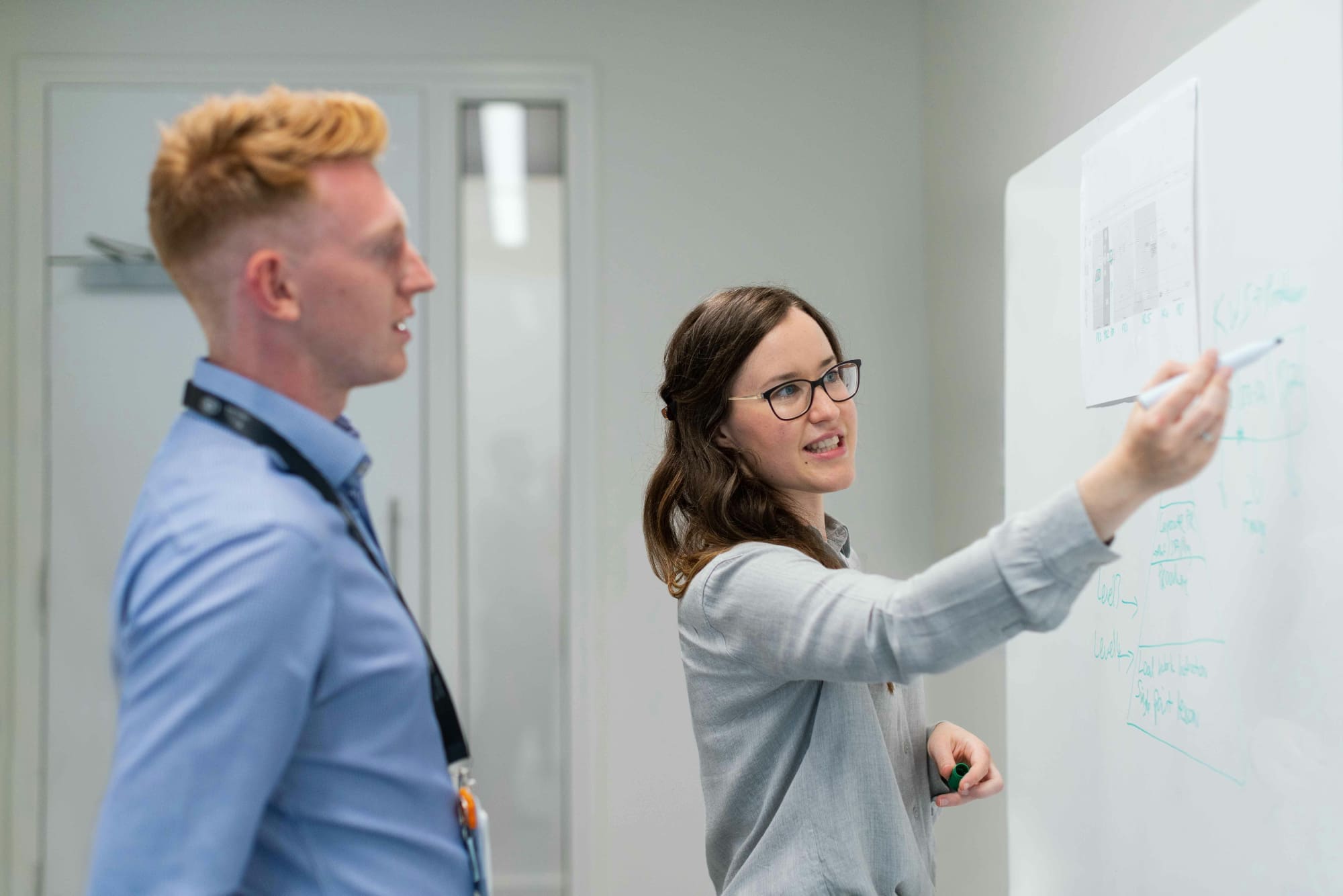 A woman explaining something to a man by pointing to a graph on a whiteboard using a marker.