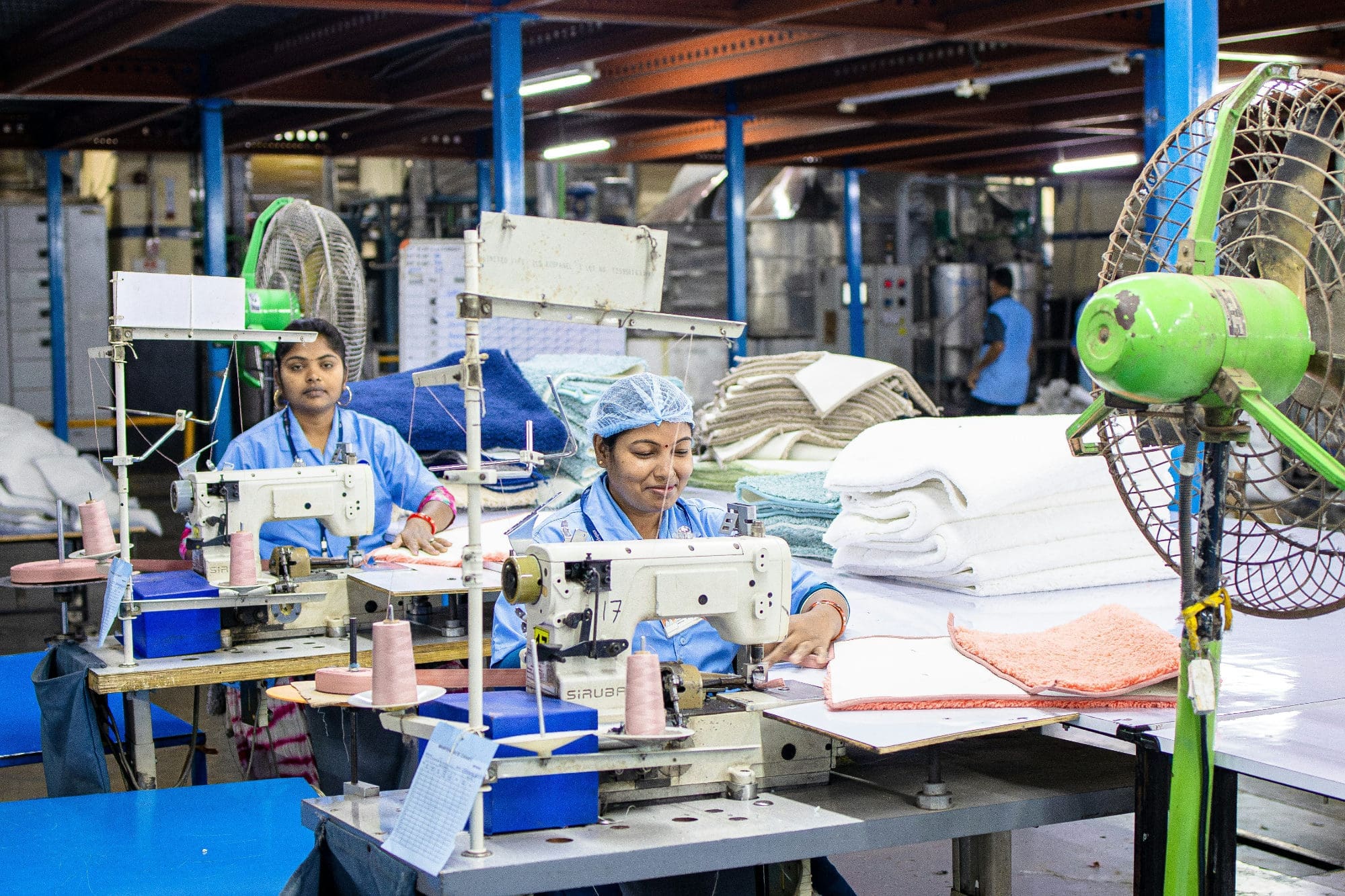 Two factory workers operating sewing machines next to a pile of garments.
