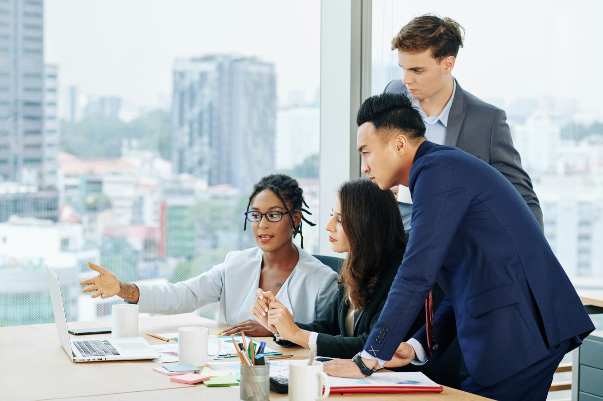 Best sustainability consultant platforms One business person pointing at laptop screen on desk with three others standing behind them looking at the screen with big office window in the background looking out onto a cityscape.