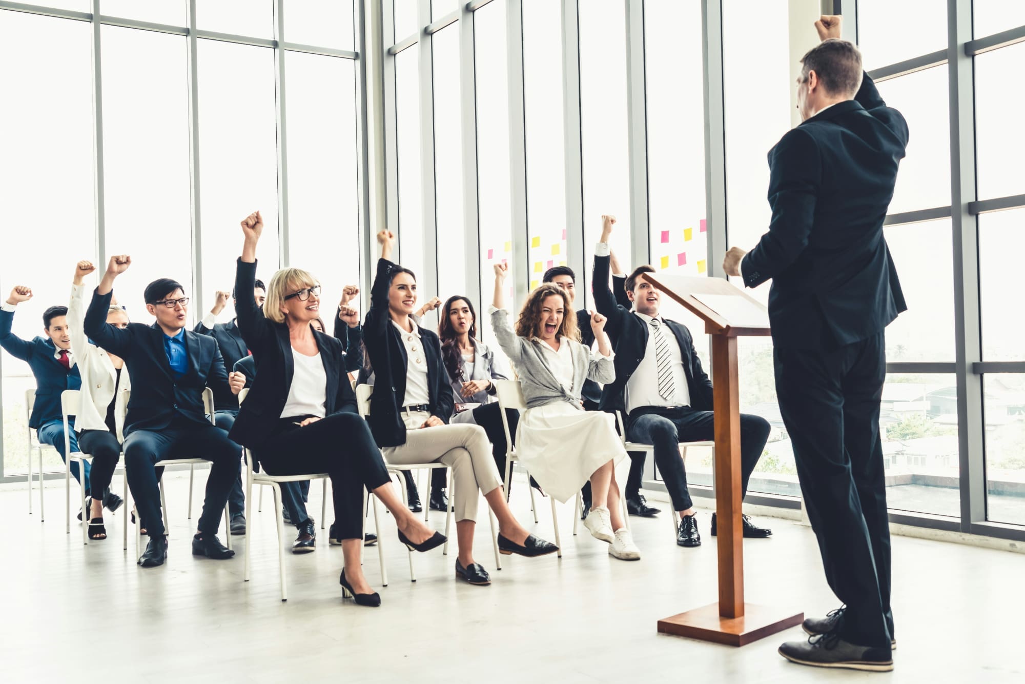 Engage stakeholders Standing business person giving presentation to other sitting business people who are raising their hands.