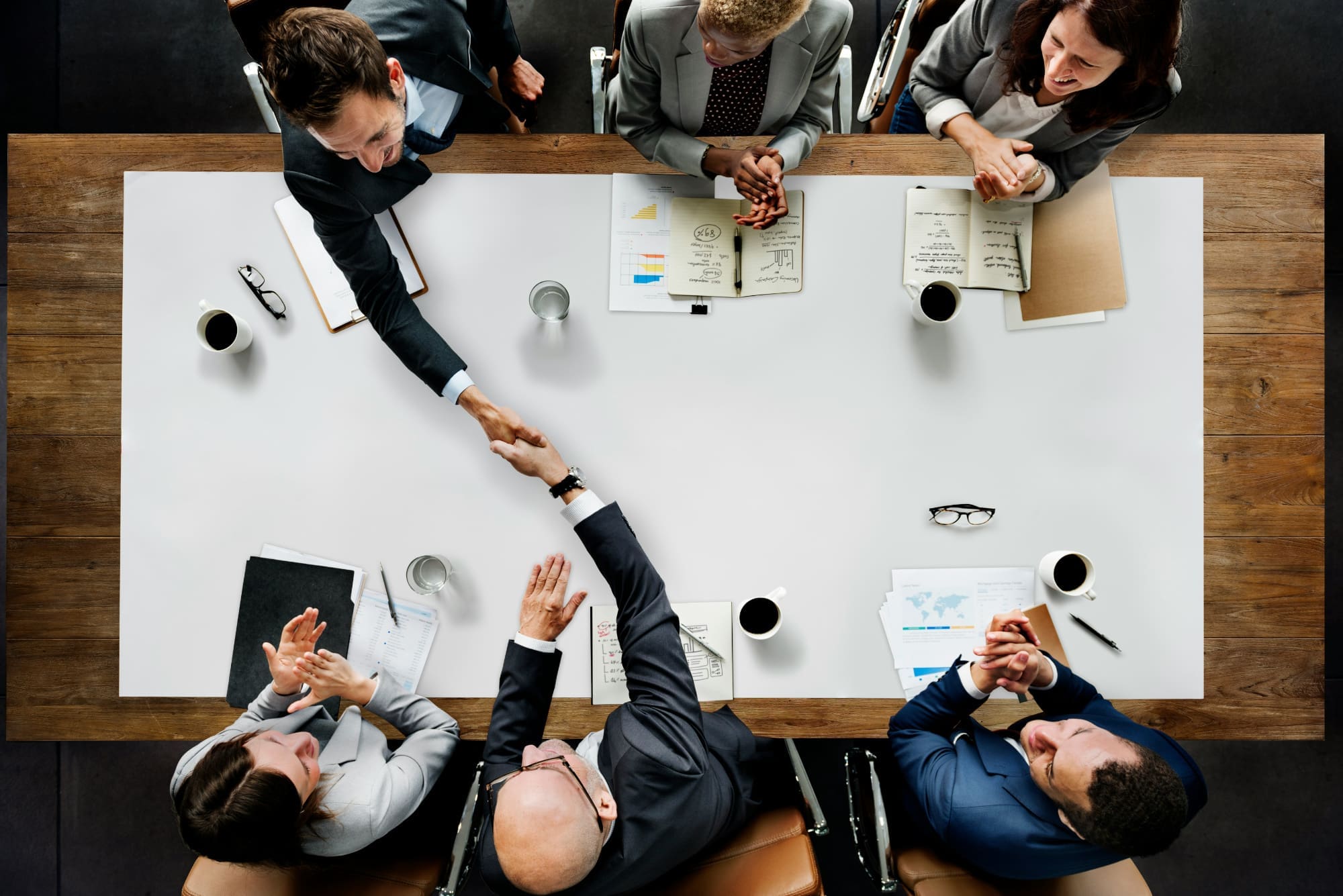 Making sustainability consultants accessible Overhead shot of three business people sitting across table from three others, with two shaking hands across table with notepads, pens, and coffee cups on it.